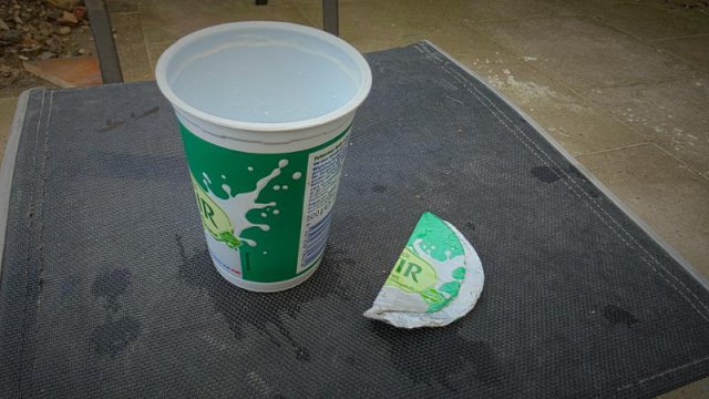 Leerer Kefirbecher und abgetrennter Metalldeckel auf einem Tisch zur Veranschaulichung der korrekten M&uuml;lltrennung.