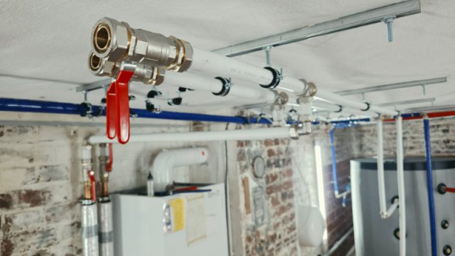 An der Decke montierte wei&szlig;e Verbundrohre mit markanten Schraubfittings und zwei roten Absperrventilen in einem Keller.