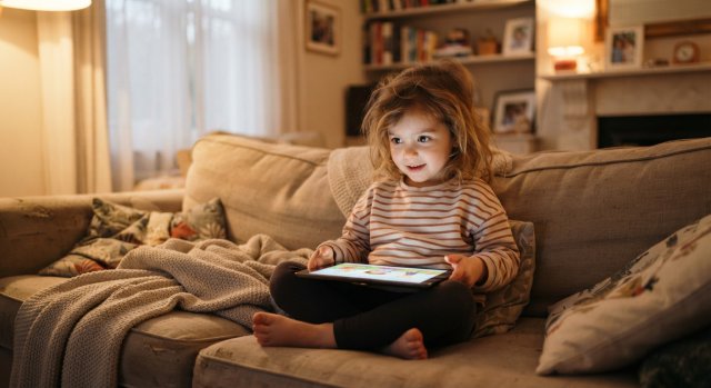 Ein Kind schaut konzentriert auf ein Tablet, w&auml;hrend im Hintergrund ein Elternteil unterst&uuml;tzend dabei sitzt.